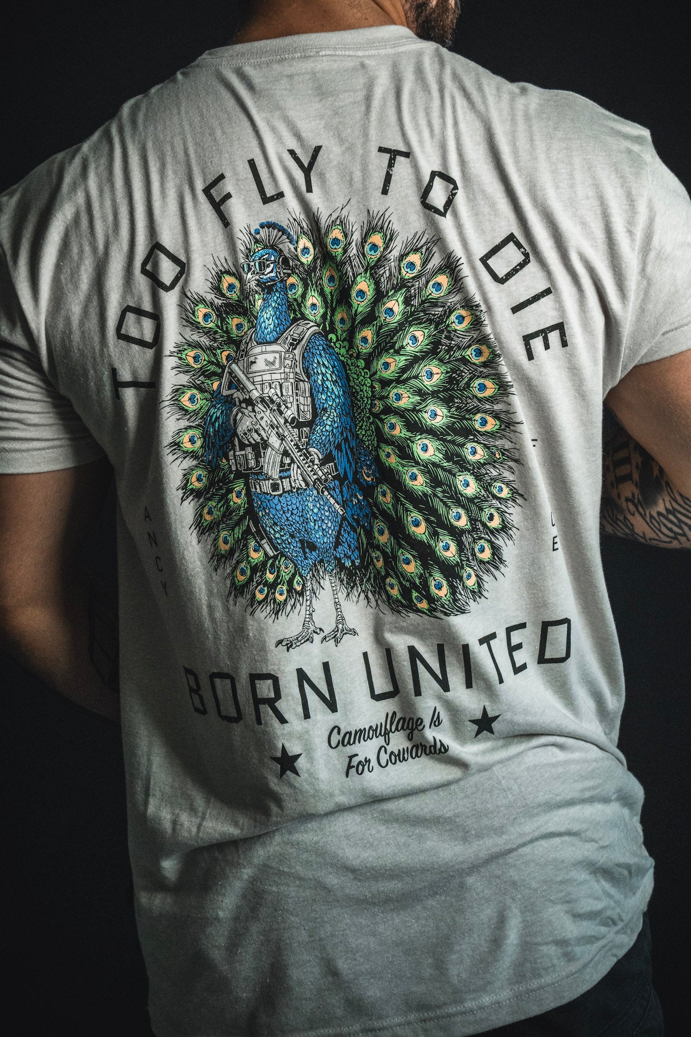 Wearing the “Too Fly To Die” T-shirt from the Born United Private Label, a person sports a bold design of a peacock holding a rifle with striking text: Too fly to die, born united, camouflage is for cowards.