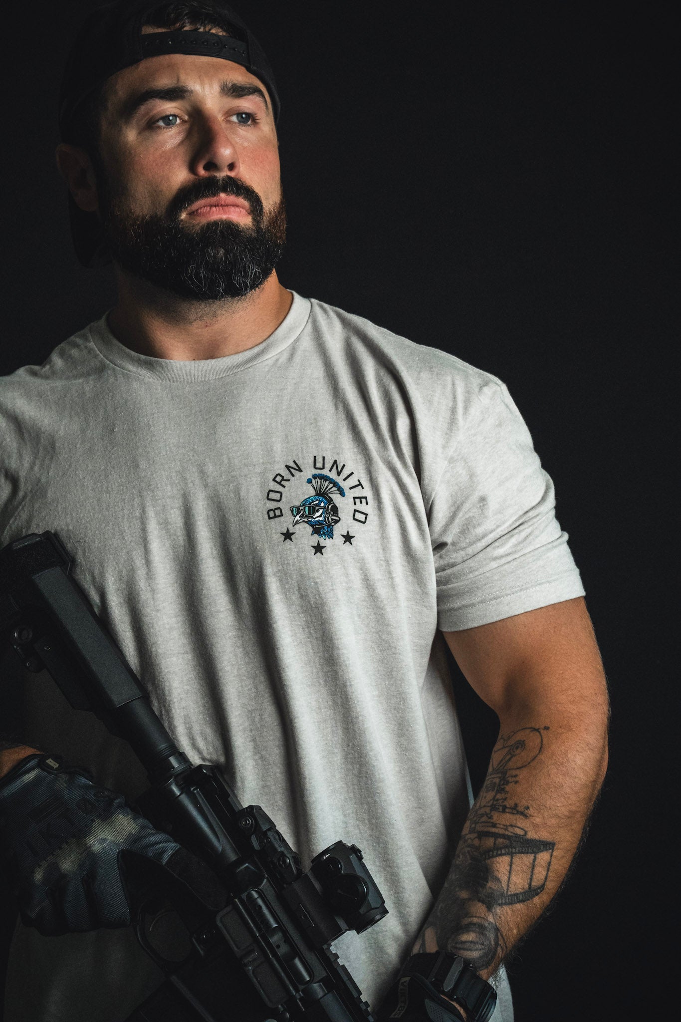 A bearded man wearing a Born United Private Label “Too Fly To Die” t-shirt and a backward cap holds a rifle against a dark background, blending seamlessly into the shadows like true Fancy Force camouflage.