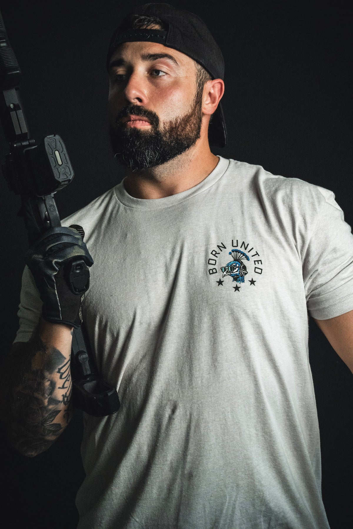 A bearded man in a cap, dressed in the white “Too Fly To Die” t-shirt from Born United Private Label with subtle camouflage accents, holds a firearm and poses confidently against a dark background—embodying the true spirit of Fancy Force.