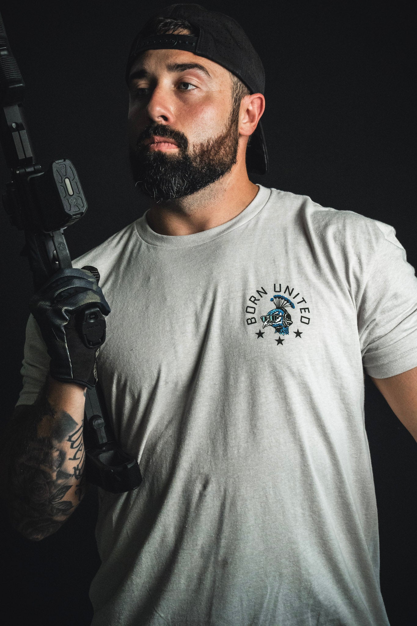A bearded man in a cap, dressed in the white “Too Fly To Die” t-shirt from Born United Private Label with subtle camouflage accents, holds a firearm and poses confidently against a dark background—embodying the true spirit of Fancy Force.