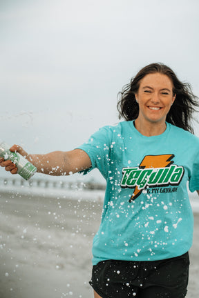 A smiling woman in a Tequila Is My Gatorade - Berry Blue shirt by Tango Charlie Apparel splashes water from a bottle on a summer beach, with bold colors and a blurred ocean and pier in the background.