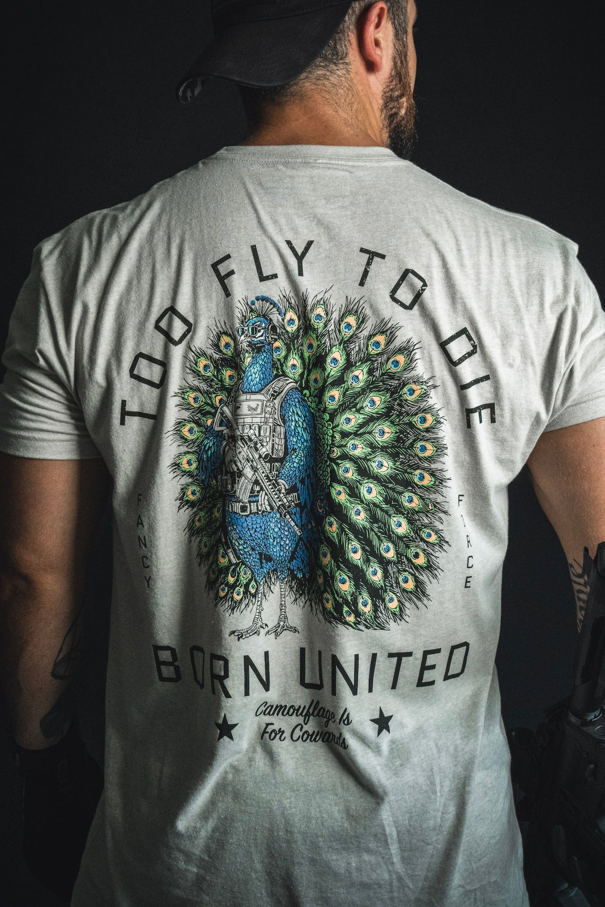 A person wearing the “Too Fly To Die” T-shirt from Born United Private Label, featuring a peacock in tactical gear and surrounded by the text “Camouflage is for cowards. Fancy Force.”.