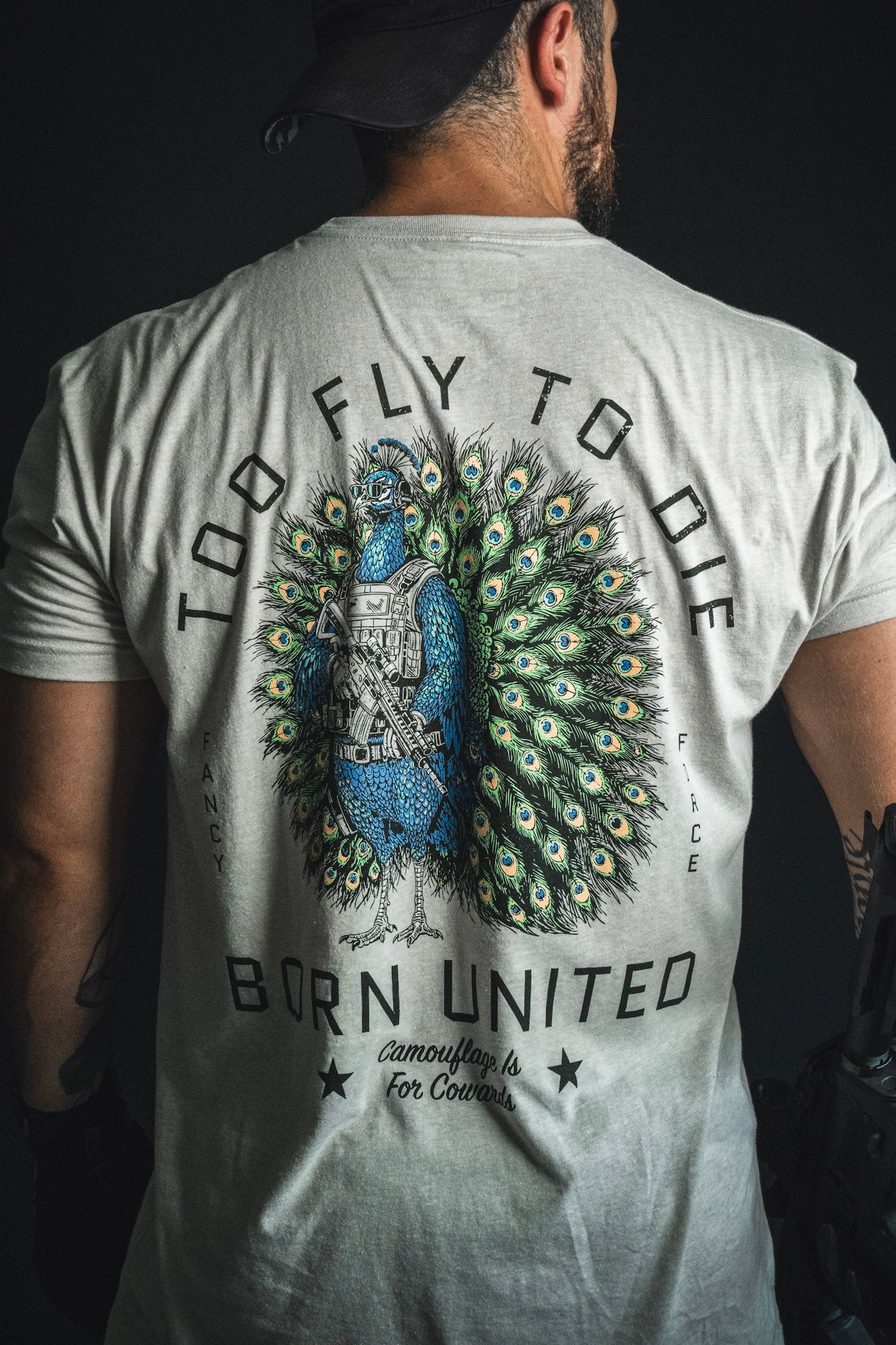 A person wearing the “Too Fly To Die” T-shirt from Born United Private Label, featuring a peacock in tactical gear and surrounded by the text “Camouflage is for cowards. Fancy Force.”.