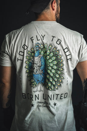 A person wearing the “Too Fly To Die” T-shirt from Born United Private Label, featuring a peacock in tactical gear and surrounded by the text “Camouflage is for cowards. Fancy Force.”.