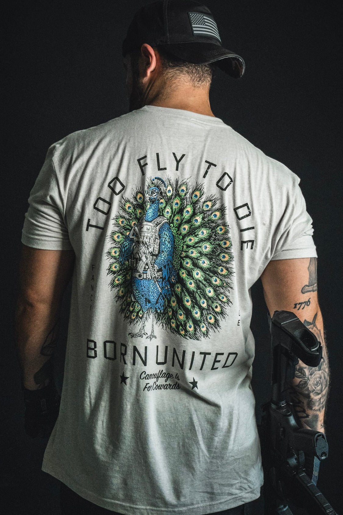 A tattooed man with a prosthetic arm sports the Born United Private Label “Too Fly To Die” shirt, featuring a striking peacock graphic and bold lettering. Facing away from the camera, he embodies confidence and style—making his statement clear without hiding behind camouflage.