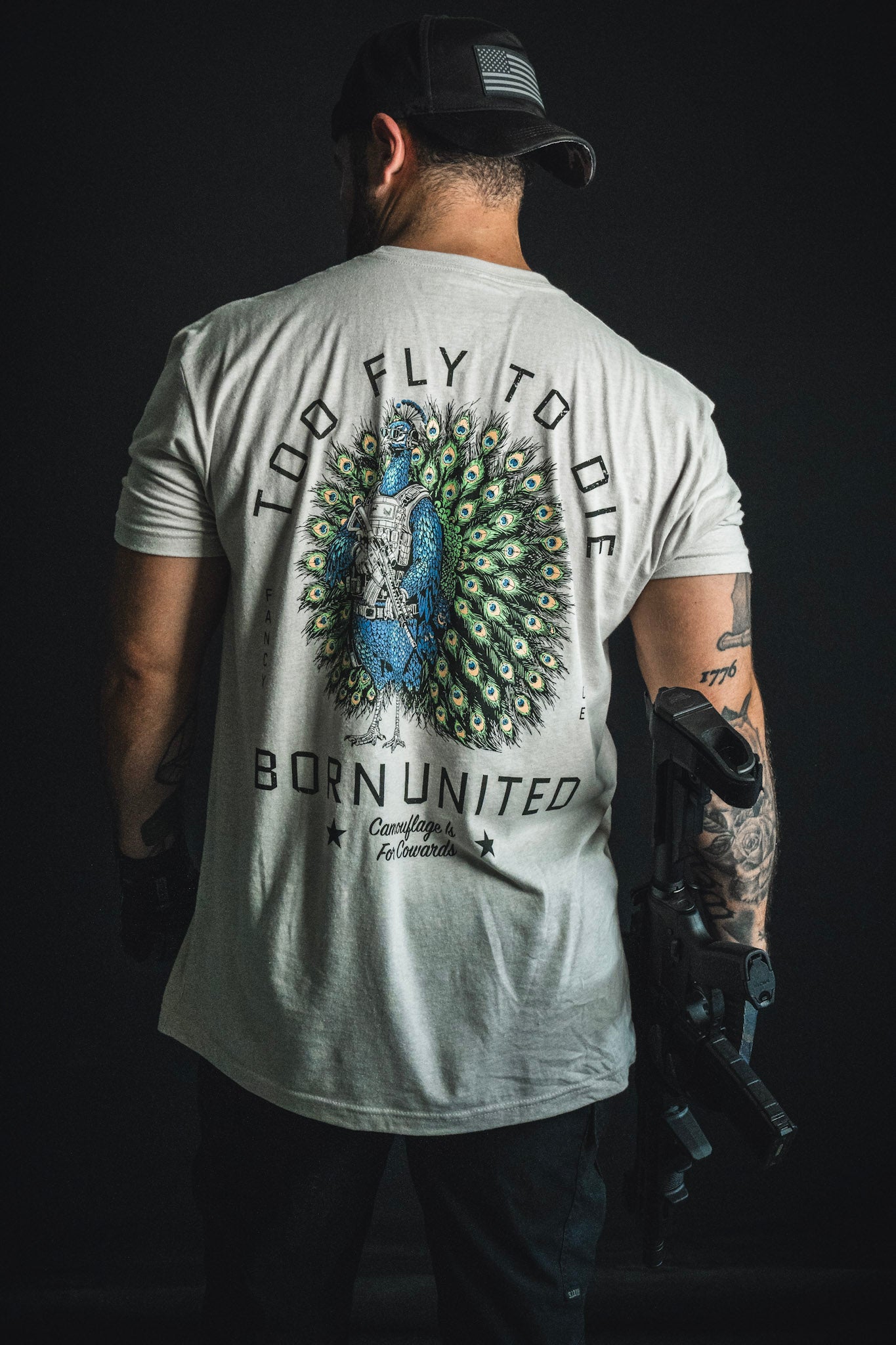 A tattooed man with a prosthetic arm sports the Born United Private Label “Too Fly To Die” shirt, featuring a striking peacock graphic and bold lettering. Facing away from the camera, he embodies confidence and style—making his statement clear without hiding behind camouflage.