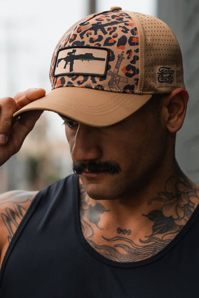 A man with a mustache and chest tattoos wears the Loaded Leopard Hydro Snapback Hat by Tango Charlie Apparel, featuring a rifle patch. He looks down, holding the brim of this beige and leopard-print hat, dressed in a black tank top.