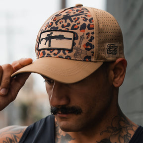 A man with a mustache adjusts the brim of his Loaded Leopard Hydro Snapback Hat by Tango Charlie Apparel, sporting a tan and leopard-print design with a rifle graphic, while standing outside near a wall.