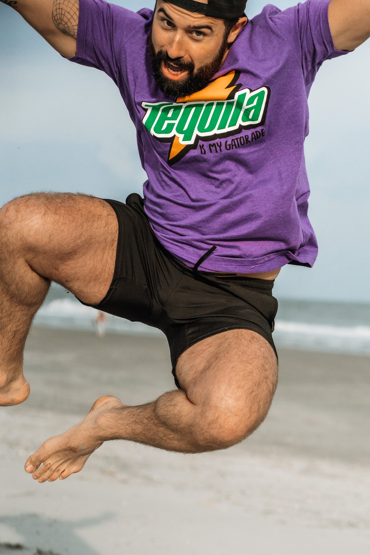 A bearded man in black shorts jumps energetically on a sandy beach, wearing a bold purple “Tequila Is My Gatorade - Purple Rush” shirt from Tango Charlie Apparel, with the ocean and sky in the background.