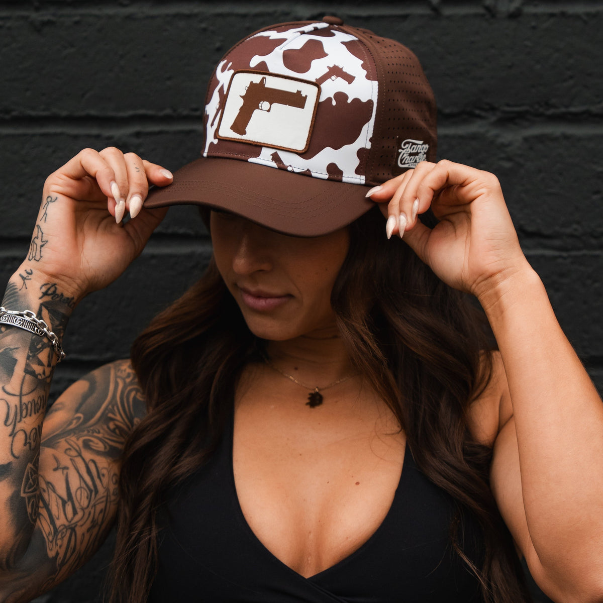 A woman with long dark hair and tattooed arms wears the Moo Moo Pew Hydro - Snapback Hat by Tango Charlie Apparel, featuring a brown camo pattern and handgun graphic, tilting the cap down with both hands against a dark brick wall.