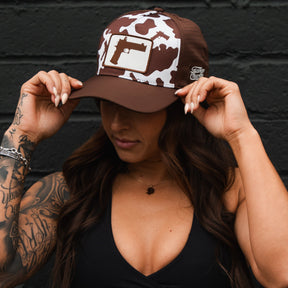 A woman with long dark hair and tattooed arms wears the Moo Moo Pew Hydro - Snapback Hat by Tango Charlie Apparel, featuring a brown camo pattern and handgun graphic, tilting the cap down with both hands against a dark brick wall.