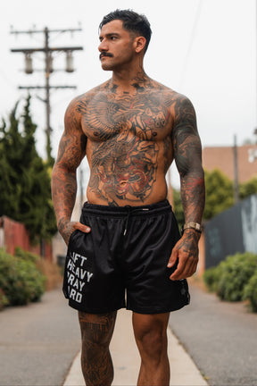 A muscular, tattooed man with a mustache walks outdoors in Tango Charlie Apparel's Men's Mesh Lifestyle Shorts | 5.5"- Lift Heavy Pray Hard Black, glancing to the side amid trees, buildings, and power lines.