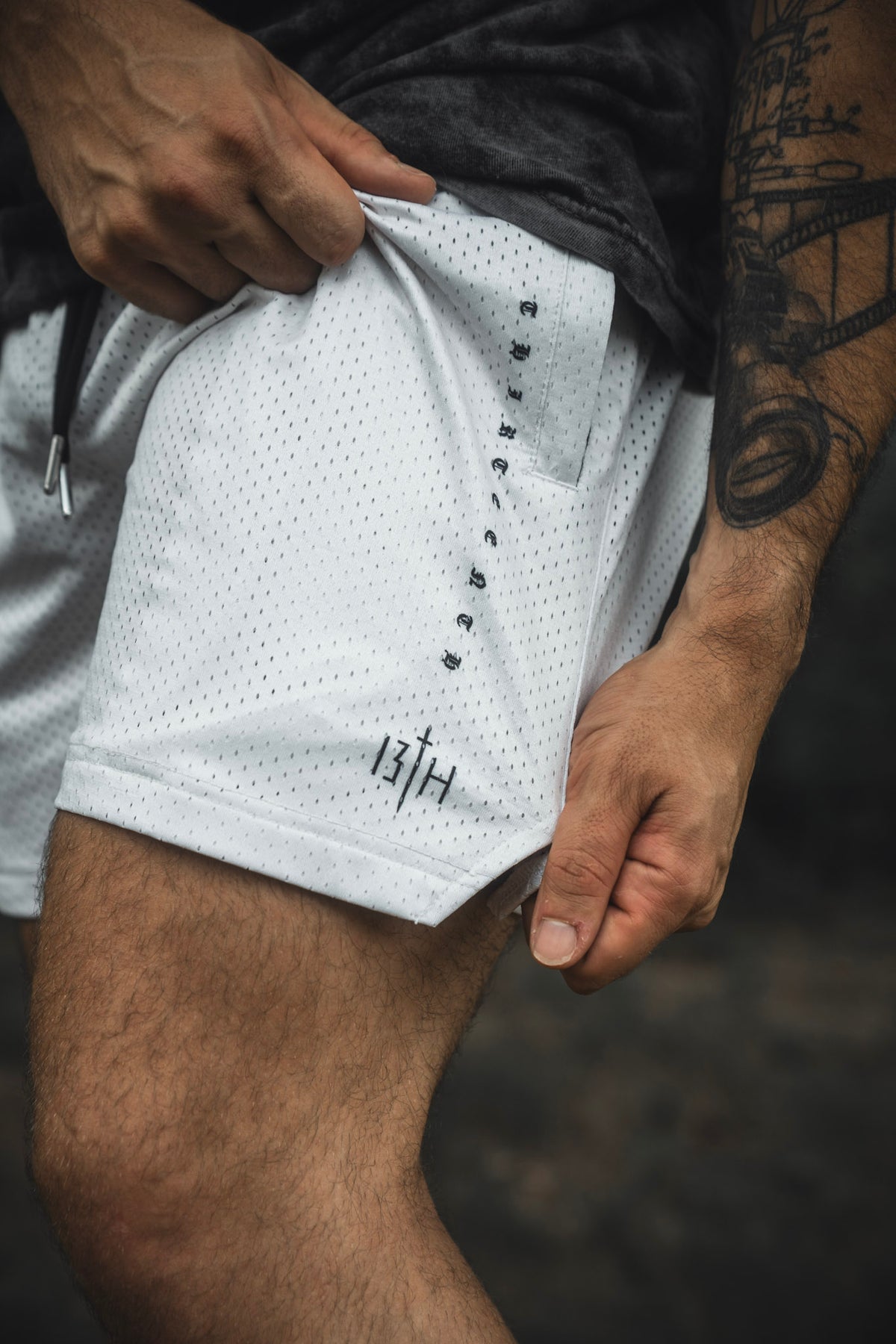 A person holds the fabric of the 13th Men’s Mesh Lifestyle Shorts | 5.5″- Christ is King Ivory, showing a tattooed arm and part of their leg; the unisex ivory mesh shorts feature small black text on the side.