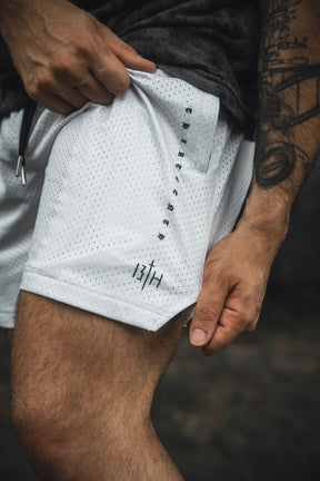 A person holds the fabric of the 13th Men’s Mesh Lifestyle Shorts | 5.5″- Christ is King Ivory, showing a tattooed arm and part of their leg; the unisex ivory mesh shorts feature small black text on the side.