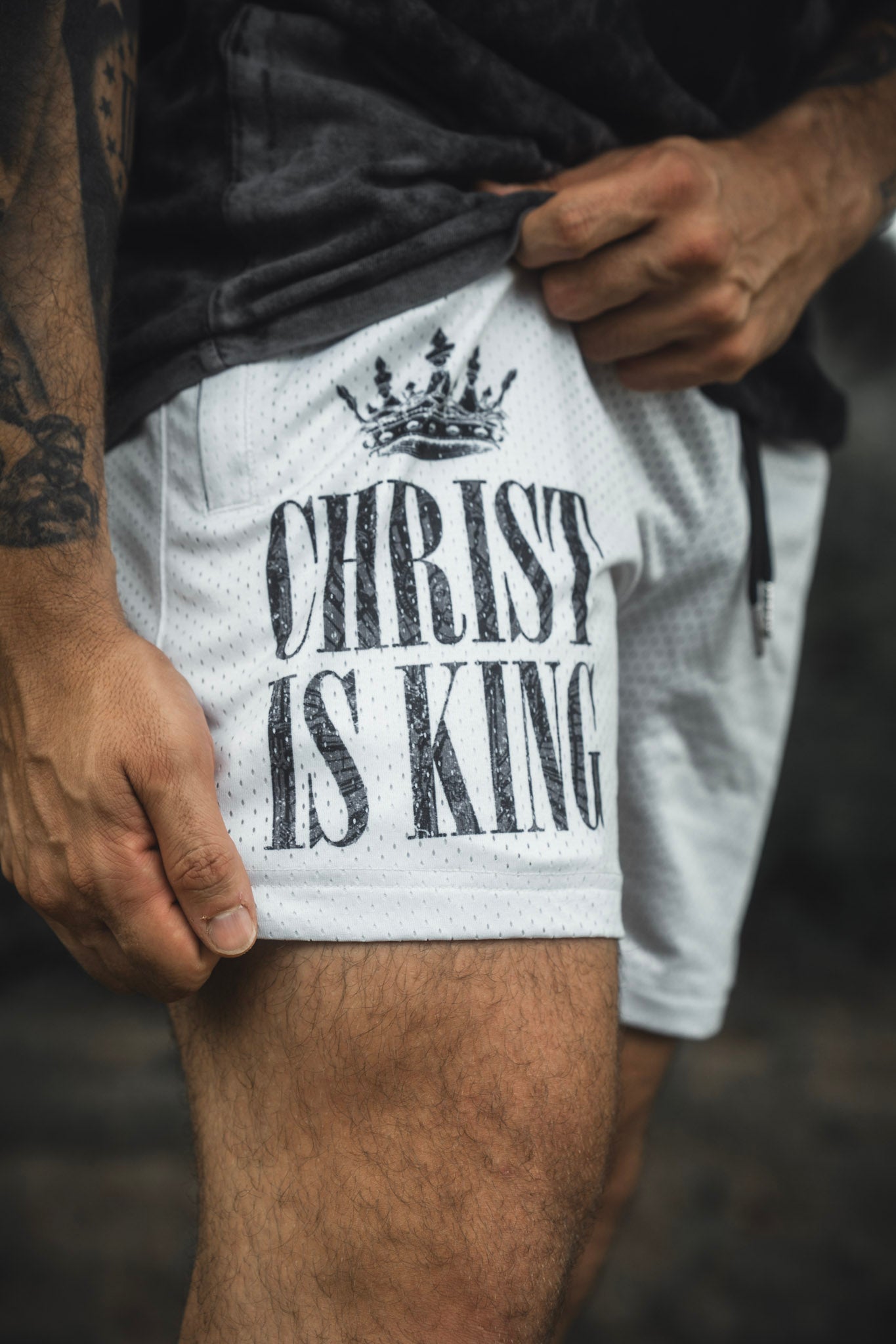 A person wears the 13th Men’s Mesh Lifestyle Shorts in Ivory, featuring a black crown graphic and “CHRIST IS KING” printed on the front.
