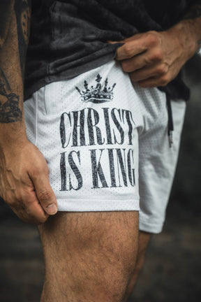 A person wears the 13th Men’s Mesh Lifestyle Shorts in Ivory, featuring a black crown graphic and “CHRIST IS KING” printed on the front.
