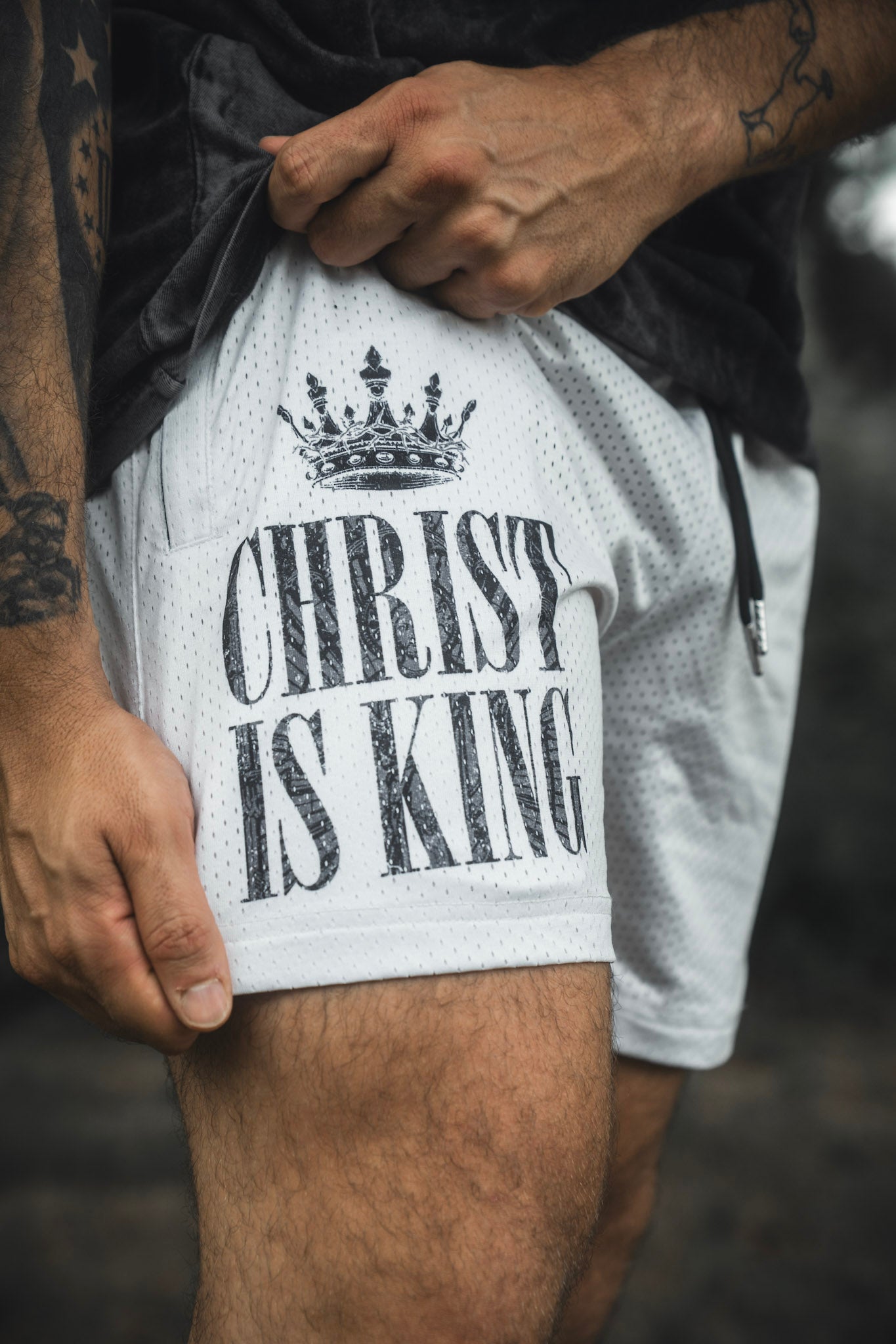 A person holds the hem of the 13th Men’s Mesh Lifestyle Shorts | 5.5″ - Christ is King Ivory, which feature a crown graphic and “Christ is King” text.