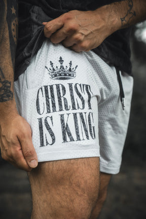 A person holds the hem of the 13th Men’s Mesh Lifestyle Shorts | 5.5″ - Christ is King Ivory, which feature a crown graphic and “Christ is King” text.