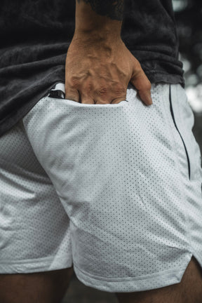 Wearing the 13th Men’s Mesh Lifestyle Shorts | 5.5″ - Christ is King Ivory, a person stands with one hand in the pocket, revealing part of their upper leg and hand.