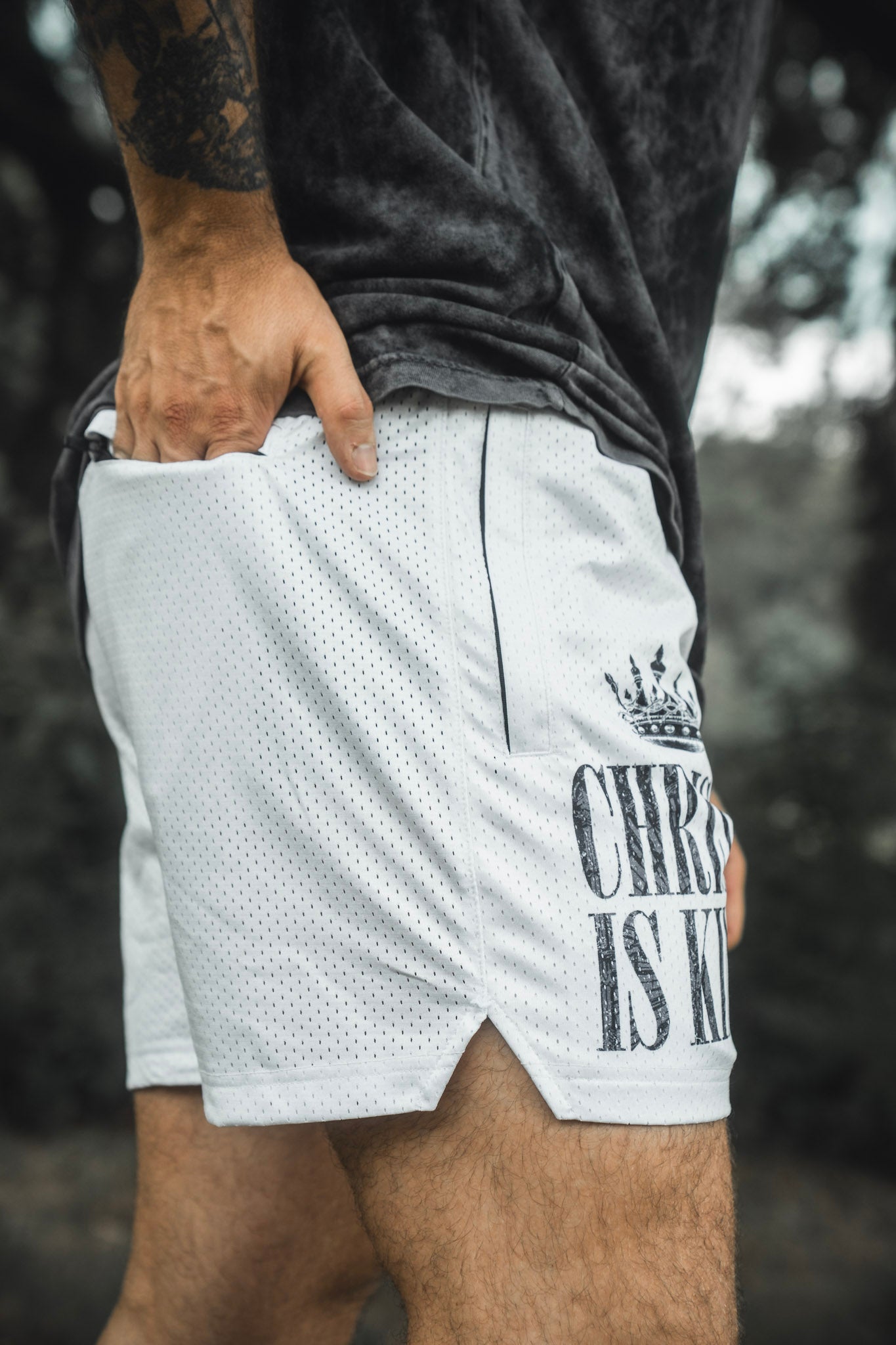 A person stands outdoors with their hand in the pocket, wearing 13th Men’s Mesh Lifestyle Shorts | 5.5″ - Christ is King Ivory, featuring a white unisex design and a “CHRIST IS KING” crown graphic printed on the left leg.