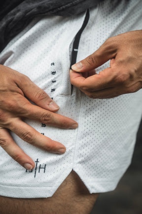 Close-up of a person unzipping a pocket on the 13th Men’s Mesh Lifestyle Shorts | 5.5″ - Christ is King Ivory, featuring white perforated mesh fabric and a small logo near the hem, highlighting these unisex performance shorts.