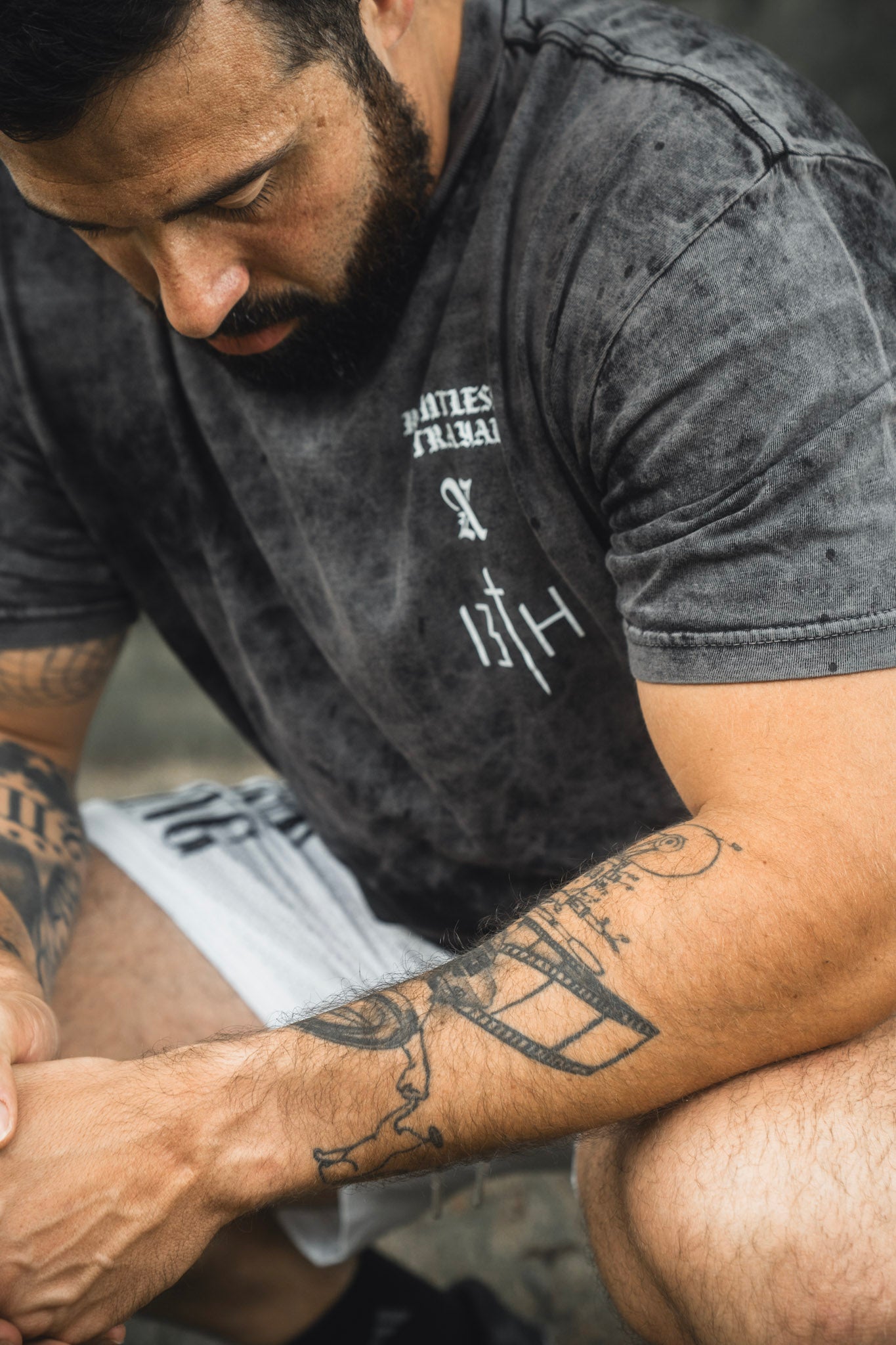 A bearded man with arm tattoos sits with his hands clasped, wearing a dark t-shirt and 13th Men’s Mesh Lifestyle Shorts | 5.5″- Christ is King Ivory.