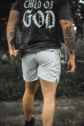 Wearing the 13th Men’s Mesh Lifestyle Shorts | 5.5″ - Christ is King Ivory, a person stands outdoors showing arm tattoos and holding up their shirt.