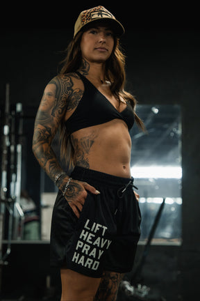 A muscular woman stands confidently in a gym wearing Tango Charlie Apparel’s Men’s Mesh Lifestyle Shorts | 5.5"- Lift Heavy Pray Hard Black, paired with a black sports bra and cap, her tattoos visible as she poses with hands in her pockets.