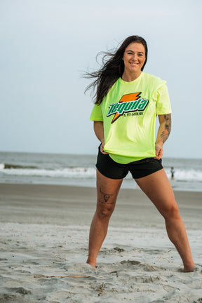 A woman with long dark hair smiles on a sandy beach near the ocean, wearing Tango Charlie Apparel’s bright yellow Tequila Is My Gatorade - Lemon Lime t-shirt and black shorts, radiating confidence as waves roll in behind her.