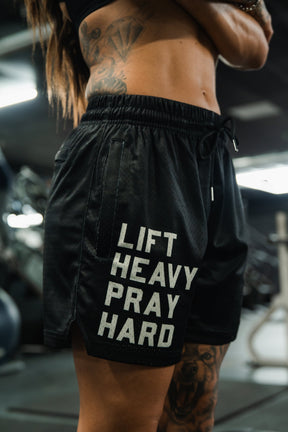A tattooed man stands in a gym, surrounded by fitness equipment, wearing Tango Charlie Apparel Men's Mesh Lifestyle Shorts | 5.5"- Lift Heavy Pray Hard Black.
