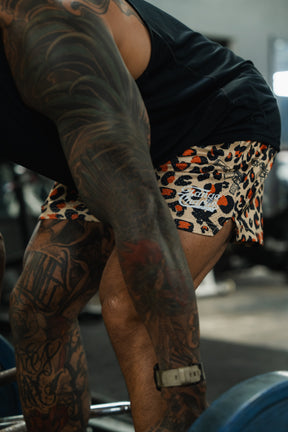 A tattooed person in a black shirt and Tango Charlie Apparel Men's Mesh Lifestyle Shorts (5.5"- Loaded Leopard) bends over to lift a barbell in a gym.