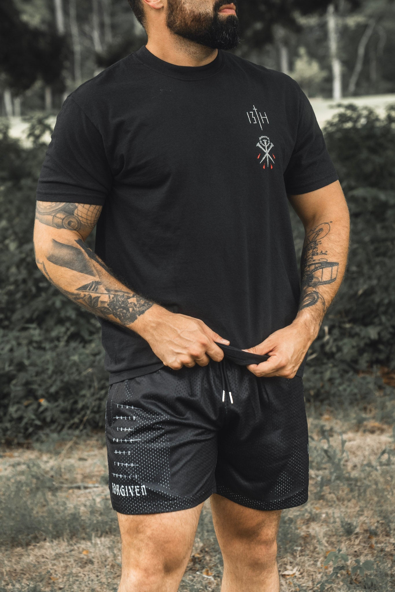 A tattooed man stands outdoors in a grassy area, wearing a black t-shirt with a small white emblem and 13th brand Men’s Mesh Lifestyle Shorts | 5.5″ - Forgiven, which feature the word “FORGIVEN” printed on the leg.