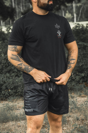 A tattooed man stands outdoors in a grassy area, wearing a black t-shirt with a small white emblem and 13th brand Men’s Mesh Lifestyle Shorts | 5.5″ - Forgiven, which feature the word “FORGIVEN” printed on the leg.