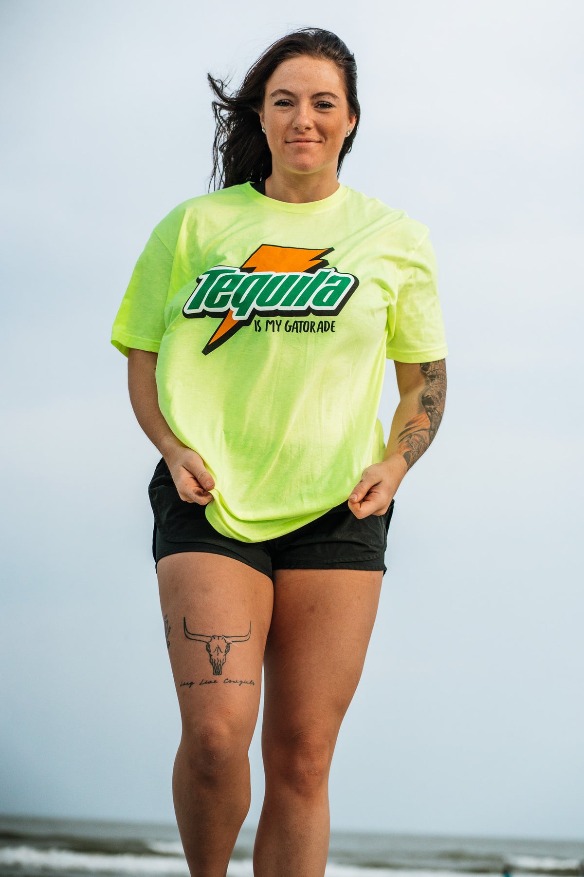 A woman stands outdoors in a lemon-lime “Tequila Is My Gatorade” T-shirt by Tango Charlie Apparel, paired with black shorts. Her long dark hair and tattoos stand out against the cloudy sky.