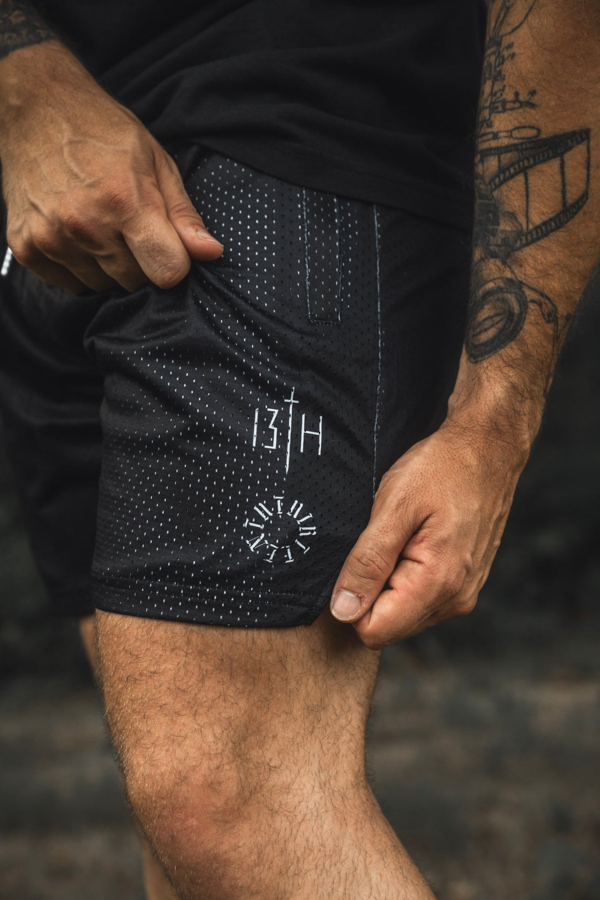 A person wearing the 13th Men’s Mesh Lifestyle Shorts | 5.5″- Forgiven, featuring black perforated fabric and compass-inspired 13H designs on the left leg, with their hand resting on the unisex shorts and tattoos visible on their right arm.