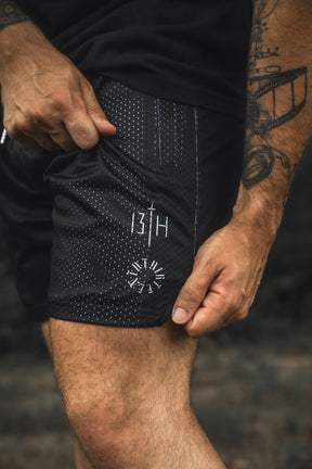 A person wearing the 13th Men’s Mesh Lifestyle Shorts | 5.5″- Forgiven, featuring black perforated fabric and compass-inspired 13H designs on the left leg, with their hand resting on the unisex shorts and tattoos visible on their right arm.