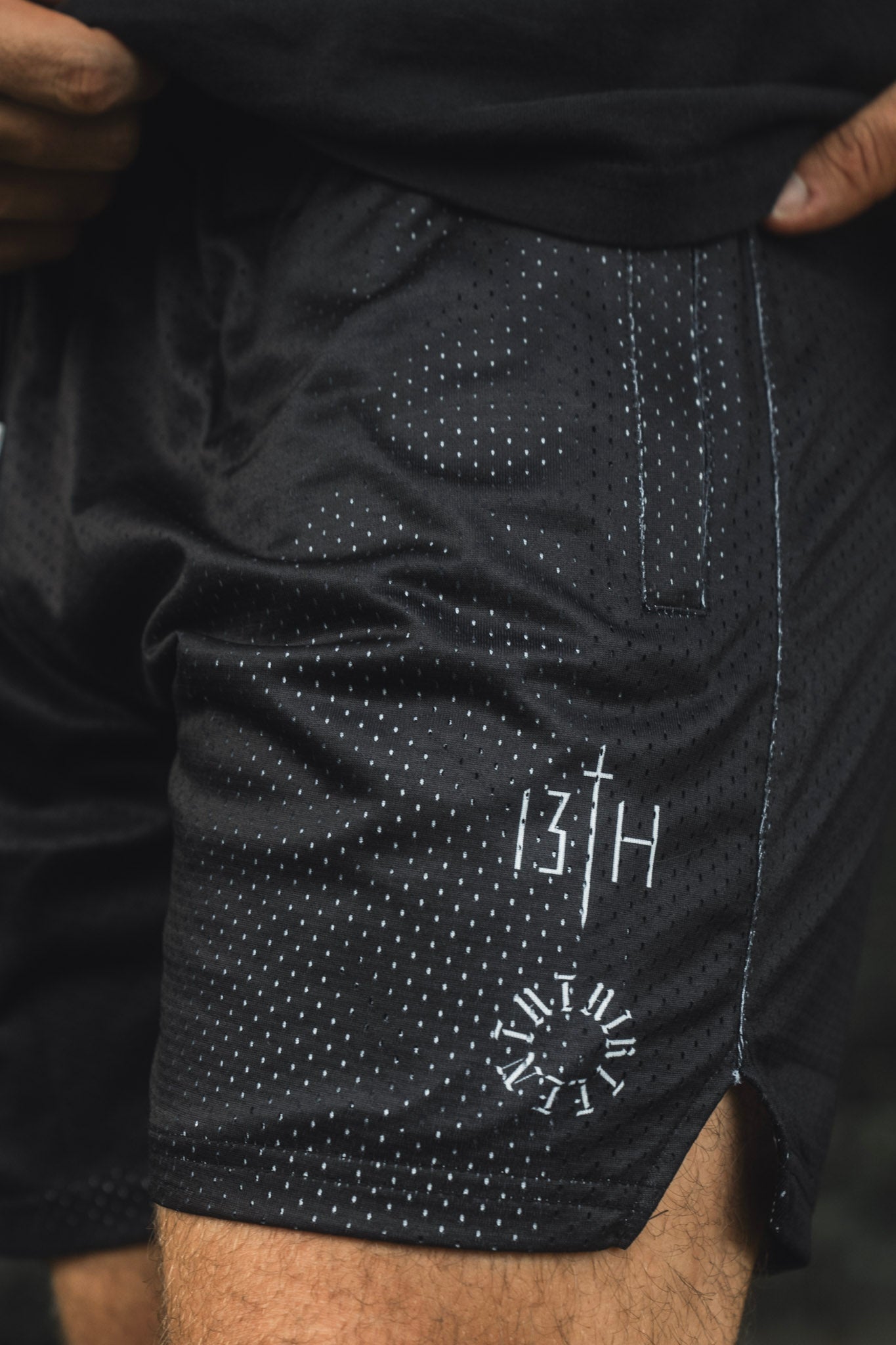 A person is wearing the Men’s Mesh Lifestyle Shorts | 5.5″ - Forgiven by 13th, featuring a black mesh fabric with a white dotted pattern, convenient pocket, and distinctive graphics—including “13TH” and a clock motif on the left leg. These unisex shorts combine comfort and style, perfect for any active lifestyle.
