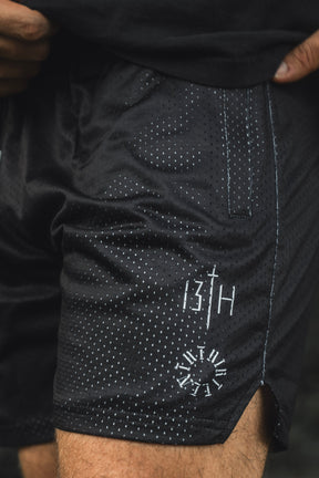 A person is wearing the Men’s Mesh Lifestyle Shorts | 5.5″ - Forgiven by 13th, featuring a black mesh fabric with a white dotted pattern, convenient pocket, and distinctive graphics—including “13TH” and a clock motif on the left leg. These unisex shorts combine comfort and style, perfect for any active lifestyle.