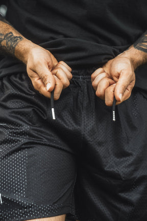 A person adjusts the drawstrings of the 13th Men’s Mesh Lifestyle Shorts | 5.5″- Forgiven in black while wearing a black shirt, with their forearms and hands visible.