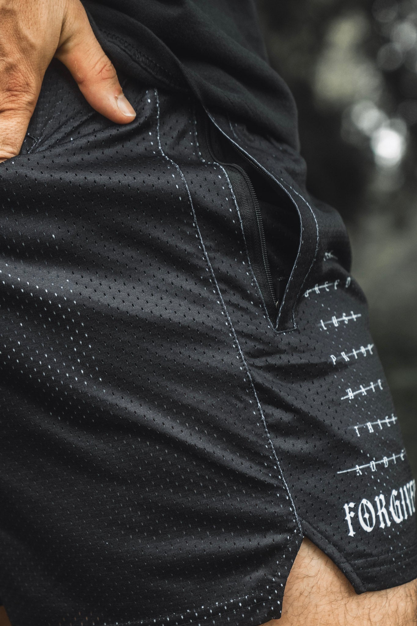 Close-up of a person wearing 13th Men’s Mesh Lifestyle Shorts | 5.5″ - Forgiven in black, featuring white stitching, with their hand resting on the waistband and partial “FORGIV” text visible on the unisex shorts fabric.