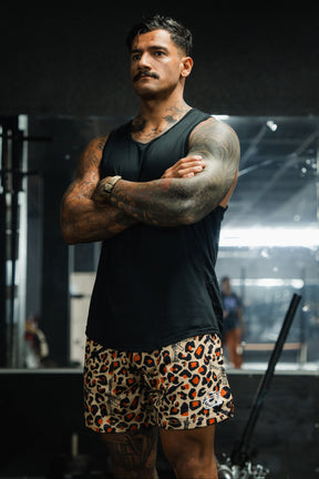 A muscular man with tattoos, wearing a black sleeveless shirt and Tango Charlie Apparel's Men's Mesh Lifestyle Shorts | 5.5"- Loaded Leopard, stands in a gym with his arms crossed, looking serious. Gym equipment is visible in the background.