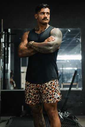 A muscular man with tattoos stands in a gym, arms crossed, wearing Tango Charlie Apparel’s Men's Mesh Lifestyle Shorts | 5.5"- Loaded Leopard and a black tank top. Gym equipment and a large mirror are visible in the background.