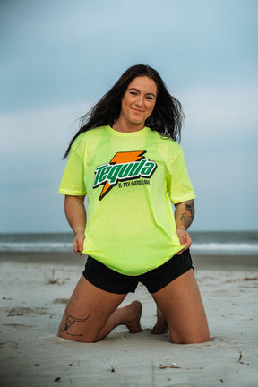 A woman with long dark hair kneels on the beach in a neon yellow Tango Charlie Apparel shirt that says Tequila Is My Gatorade - Lemon Lime and black shorts, with bold colors standing out against the ocean and cloudy sky behind her.