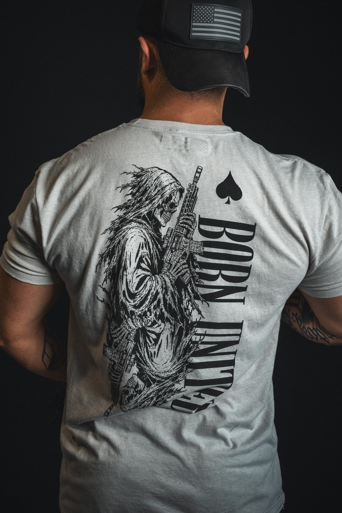A person facing away from the camera wears the Born United Private Label’s Death Card tee in light gray, featuring a skeleton with a gun and “BORN UNLUCKY” emblazoned on the back, channeling a dark aesthetic.