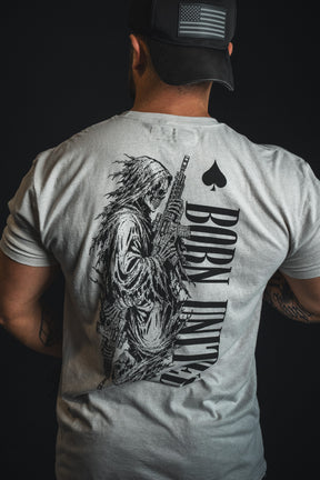 A person facing away from the camera wears the Born United Private Label’s Death Card tee in light gray, featuring a skeleton with a gun and “BORN UNLUCKY” emblazoned on the back, channeling a dark aesthetic.