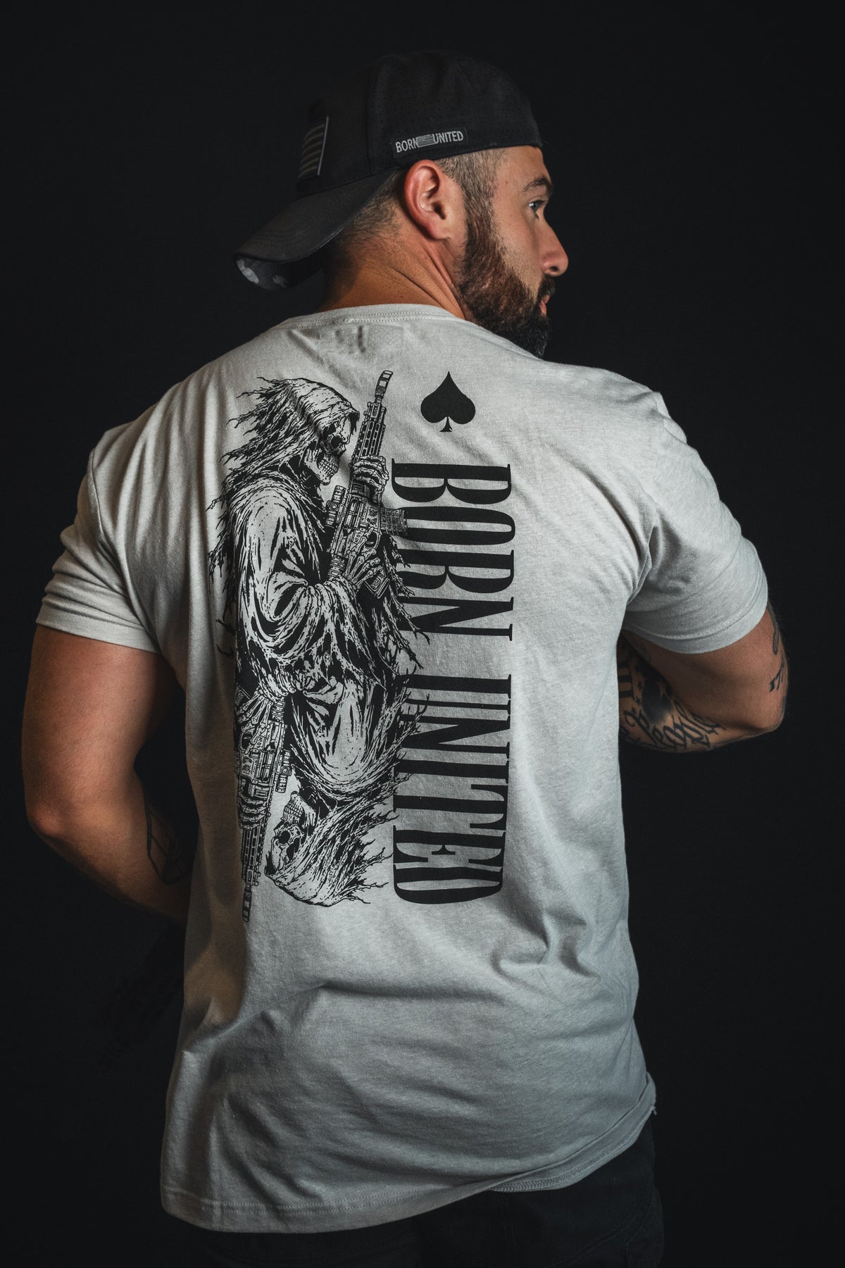 A man wearing the Born United Private Label “Death Card” T-shirt—a light gray tee featuring a bold graphic of a grim reaper wielding a rifle and the statement “BORN UNDEAD” across the back—faces away with a black cap turned backward, capturing striking dark aesthetics and wartime symbolism.