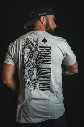 A man wearing the Born United Private Label “Death Card” T-shirt—a light gray tee featuring a bold graphic of a grim reaper wielding a rifle and the statement “BORN UNDEAD” across the back—faces away with a black cap turned backward, capturing striking dark aesthetics and wartime symbolism.