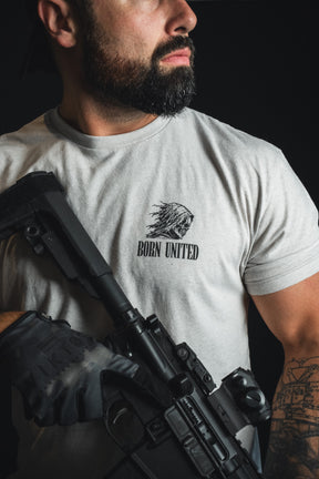 A bearded man with a tattooed arm holds a rifle while wearing the Born United Private Label “Death Card” tee, which features wartime symbolism on the chest. He is photographed against a dark background that highlights the shirt’s moody, dramatic aesthetics.