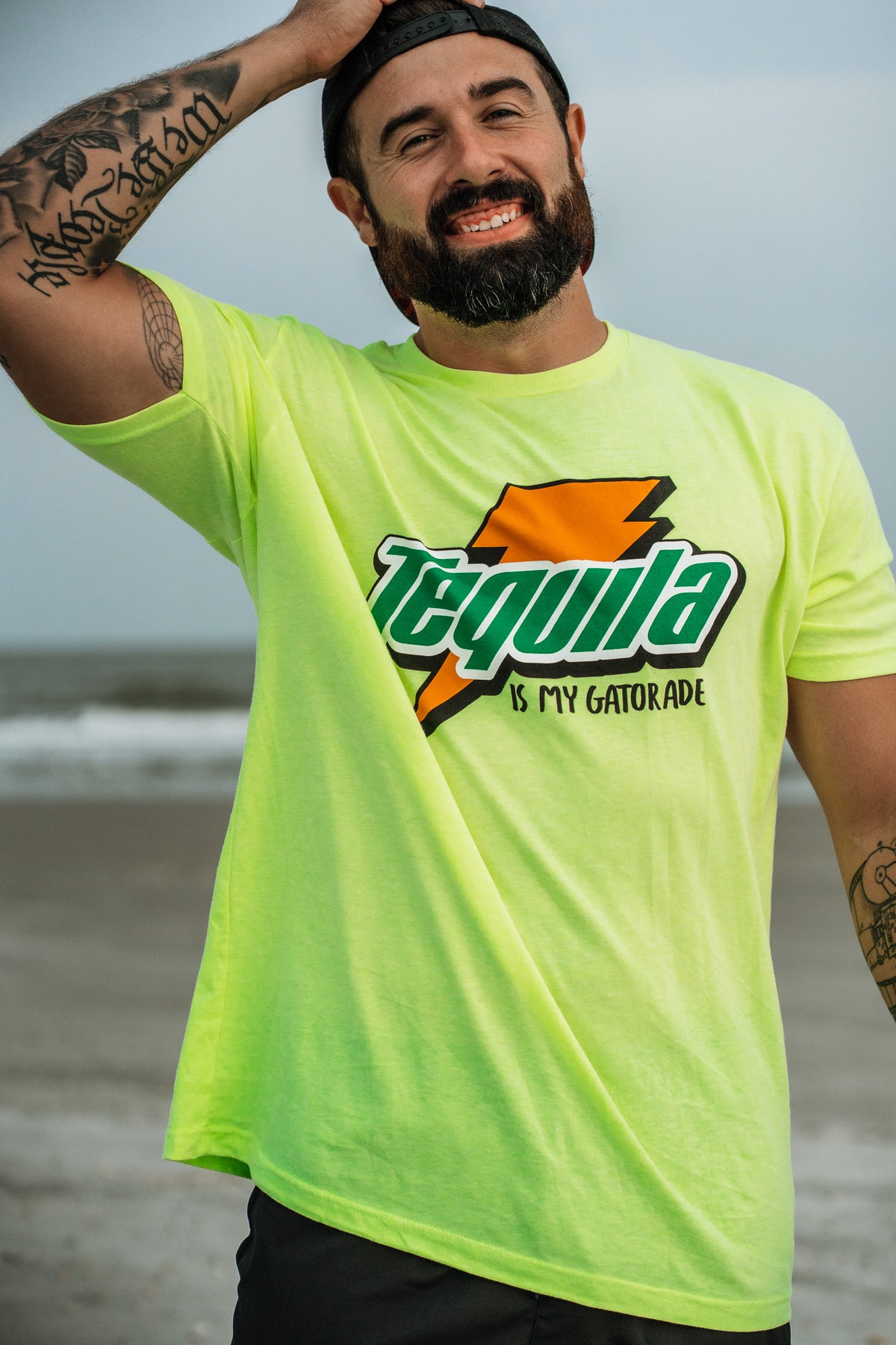 A smiling man on the beach wears a neon yellow Tango Charlie Apparel Tequila Is My Gatorade - Lemon Lime T-shirt with a lightning bolt, tattoos, beard, and backwards cap, touching his head with one hand.