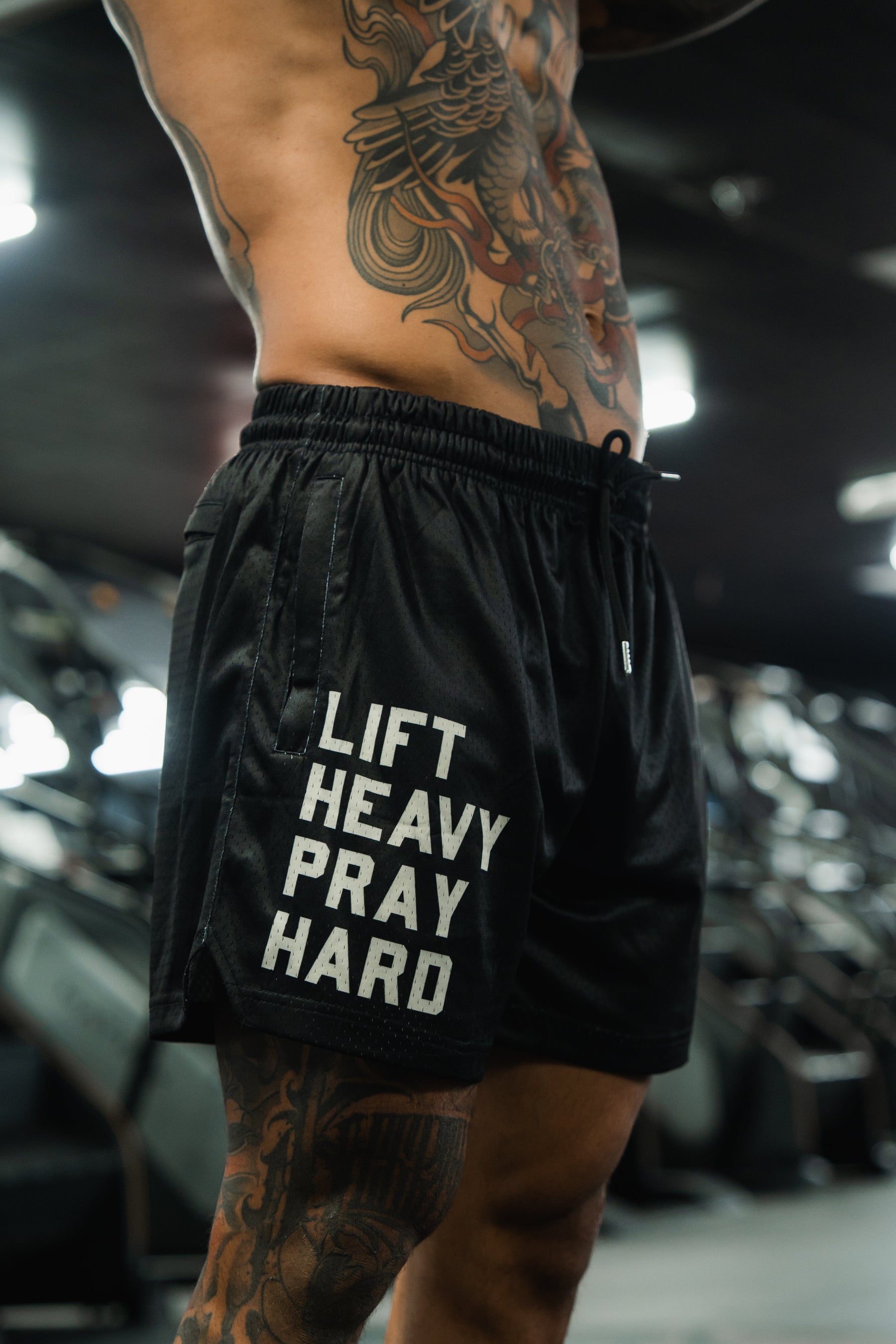 A muscular tattooed man stands in the gym, sporting Tango Charlie Apparel’s Men's Mesh Lifestyle Shorts | 5.5"- Lift Heavy Pray Hard Black, with bold slogan visible. The photo focuses on his torso and shorts, emphasizing a strong fitness lifestyle.